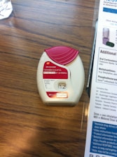 A medication inhaler labeled 'Anoro Ellipta' is placed on a wooden surface. The inhaler has a red and white design with clear dosage instructions and a counter displaying the number '12'. Beside it, part of a paper or booklet with informational text is visible.