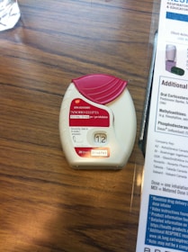 A medication inhaler labeled 'Anoro Ellipta' is placed on a wooden surface. The inhaler has a red and white design with clear dosage instructions and a counter displaying the number '12'. Beside it, part of a paper or booklet with informational text is visible.