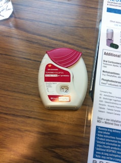 A medication inhaler labeled 'Anoro Ellipta' is placed on a wooden surface. The inhaler has a red and white design with clear dosage instructions and a counter displaying the number '12'. Beside it, part of a paper or booklet with informational text is visible.