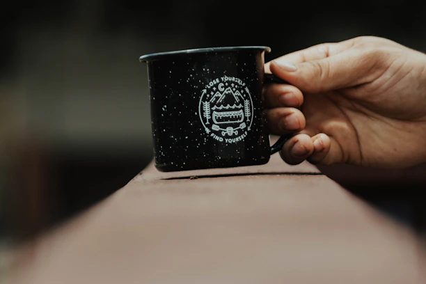 A rugged enamel mug resting on a weathered wooden table beside a glowing campfire under a misty river morning.