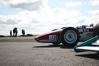 A vintage race car gleaming under sunlight, parked on a racetrack with blurred spectators in the background