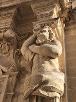 A detailed stone sculpture of a male figure is integrated into a classical architectural structure. The sculpture depicts a muscular figure with curly hair, appearing to hold up a section of the building above. The stone surface reveals intricate carvings and weathered textures, suggesting historical significance.