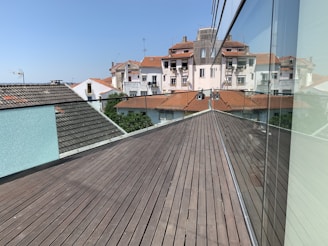 A modern rooftop terrace with wooden decking extends towards older-style apartment buildings with red tile roofs. The clear blue sky enhances the brightness of the day. A reflective glass wall runs along the terrace, mirroring the architectural surroundings.
