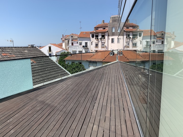 A modern rooftop terrace with wooden decking extends towards older-style apartment buildings with red tile roofs. The clear blue sky enhances the brightness of the day. A reflective glass wall runs along the terrace, mirroring the architectural surroundings.