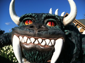 A large, fierce-looking statue with a monstrous appearance, featuring red eyes, large horns, sharp white teeth, and a dark green, textured body. The background includes some greenery and a building with a sloped roof.