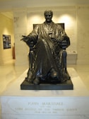 A bronze statue of a seated figure wearing a robe is prominently displayed indoors. The figure holds one hand slightly extended and the other resting on its lap, seated on a carved chair. The pedestal is inscribed with the name John Marshall along with details of his role and tenure as Chief Justice. The setting appears to be a museum or gallery with inscriptions on the wall behind the statue.