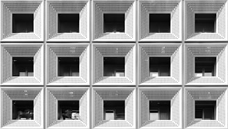 A grid of square-shaped, perforated metallic panels form the exterior facade of a building. Each panel features an inset window with a shaded interior, creating a pattern of repetitive geometric shapes. The monochrome color palette enhances the modern, industrial aesthetic.