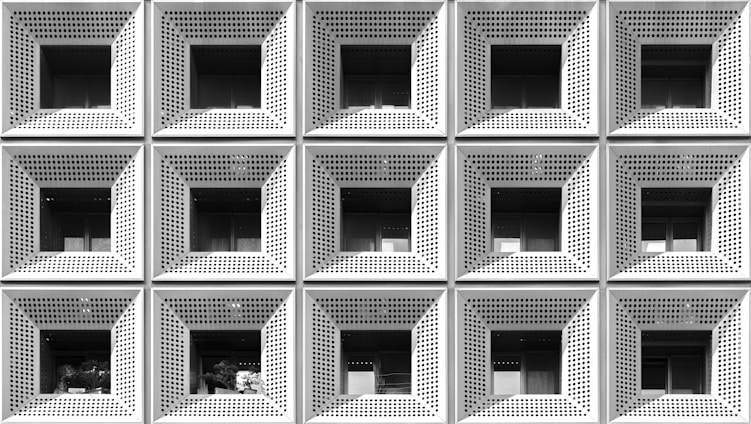 A grid of square-shaped, perforated metallic panels form the exterior facade of a building. Each panel features an inset window with a shaded interior, creating a pattern of repetitive geometric shapes. The monochrome color palette enhances the modern, industrial aesthetic.
