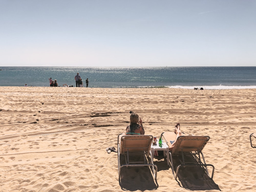 two women on brown lounger chairs beside seashore during daytime