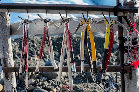 Several ice axes and climbing gear hang on a wooden rack. The axes have bright red and yellow handles with metallic tips, set against a rocky and snowy background.