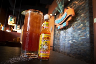 A large glass filled with a dark, iced beverage is garnished with a lime slice and salt around the rim. The glass is positioned on a table next to a bottle of Cholula hot sauce. In the background, a blurred, warm-toned restaurant environment is visible, with a textured stone wall and hanging light fixtures.