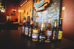 Several beer bottles are arranged on a table in a dimly lit restaurant or bar setting. The labels on the bottles are distinct, representing various brands including Corona, Modelo, Tecate, and others. The background features warm lighting and a stone wall with an orange sign visible.