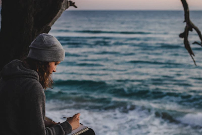 A smiling woman journaling by the sea, wrapped in a warm cream shawl.