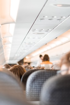 The interior of an airplane cabin is visible, focusing on the overhead compartments and rows of passenger seats. People are seated, and a flight attendant is seen standing in the aisle, wearing a uniform with orange accents.