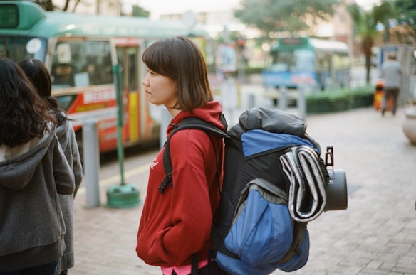 A young woman with a short haircut stands outdoors with a large blue and black backpack that includes a rolled-up mat and what might be a camera tripod attached to it. She is wearing a red hoodie and appears to be waiting at a bus station with blurred public transport buses visible in the background. Other people are present, with one wearing a gray hoodie, mostly seen from behind.