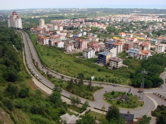 Aerial view of a suburban area featuring a curving road surrounded by lush greenery. Mid-rise residential buildings with diverse architectural styles are clustered together, with some larger complexes in the background. The landscape includes well-maintained lawns and scattered trees, providing a balance of urban and natural elements.