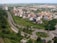 Aerial view of a suburban area featuring a curving road surrounded by lush greenery. Mid-rise residential buildings with diverse architectural styles are clustered together, with some larger complexes in the background. The landscape includes well-maintained lawns and scattered trees, providing a balance of urban and natural elements.