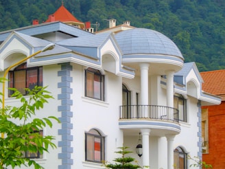 A large, elegant house with a distinctive architectural style featuring a dome-shaped roof and multiple columns. The building has a white exterior with blue-gray trim and several large windows. Surrounding the house are leafy trees and a mountainous backdrop, contributing to a serene and picturesque setting.