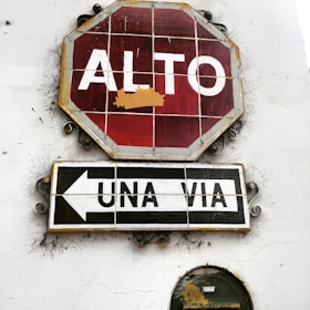 A rustic stop sign with Spanish text 'ALTO' is mounted on a white wall. Below it, a sign reading 'UNA VIA' with an arrow pointing left indicates a one-way street. The signs appear weathered and aged, with visible cracks and peeling paint.