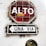 A rustic stop sign with Spanish text 'ALTO' is mounted on a white wall. Below it, a sign reading 'UNA VIA' with an arrow pointing left indicates a one-way street. The signs appear weathered and aged, with visible cracks and peeling paint.