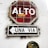 A rustic stop sign with Spanish text 'ALTO' is mounted on a white wall. Below it, a sign reading 'UNA VIA' with an arrow pointing left indicates a one-way street. The signs appear weathered and aged, with visible cracks and peeling paint.