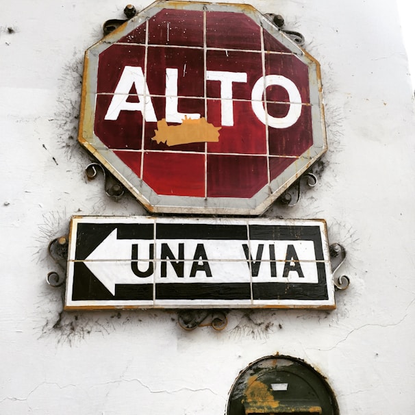 A rustic stop sign with Spanish text 'ALTO' is mounted on a white wall. Below it, a sign reading 'UNA VIA' with an arrow pointing left indicates a one-way street. The signs appear weathered and aged, with visible cracks and peeling paint.