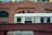 A silver train car is moving through an urban area, partially covered by a rusty, riveted steel bridge. The train has a handicap sign next to the door. A backdrop of a brick building with arched windows provides an architectural contrast.