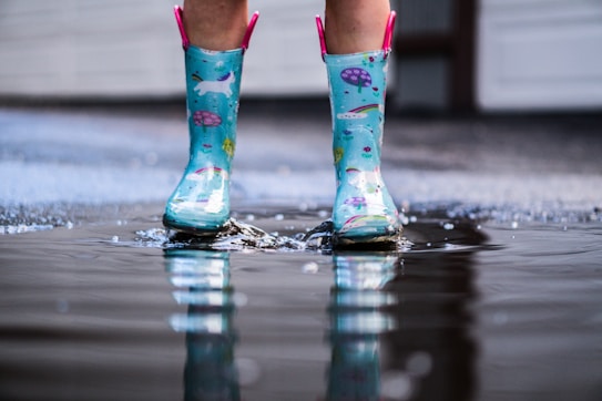 A pair of brightly colored rain boots with a playful design stands in a puddle on a wet surface. The boots appear to have a blue base with multicolored patterns, including rainbows and animals. Water splashes around as the person wearing the boots steps, creating small ripples in the puddle.