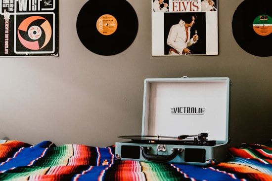A record player sits open on a colorful, striped blanket with vinyl records hanging on the wall. The wall displays records, including one featuring an iconic musician in a white suit. The room has a vintage and retro aesthetic.