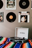 A record player sits on a colorful, striped blanket. Vinyl records are displayed on the wall featuring album covers such as Elvis. The atmosphere suggests a retro or vintage theme with a focus on music.