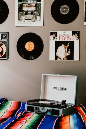 A record player sits on a colorful, striped blanket. Vinyl records are displayed on the wall featuring album covers such as Elvis. The atmosphere suggests a retro or vintage theme with a focus on music.