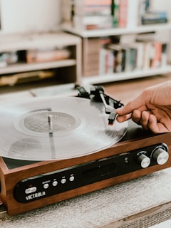 A hand carefully places the needle onto a vinyl record on a wooden turntable. The setting appears to be a cozy room with blurred bookshelves in the background, suggesting a calm and nostalgic atmosphere.
