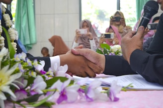 Two individuals are shaking hands over a table adorned with flowers and paperwork. A person holds a microphone nearby, while in the background, several people are taking photos with smartphones. The setting appears formal and celebratory.