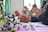 Two individuals are shaking hands over a table adorned with flowers and paperwork. A person holds a microphone nearby, while in the background, several people are taking photos with smartphones. The setting appears formal and celebratory.
