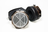 A pair of retro-style headphones with wooden casings, metal grills, and black padded ear cups lies on a white surface. The headphones feature hexagonal ear cups with a unique lattice design and a visible logo.