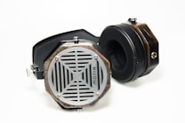 A pair of retro-style headphones with wooden casings, metal grills, and black padded ear cups lies on a white surface. The headphones feature hexagonal ear cups with a unique lattice design and a visible logo.