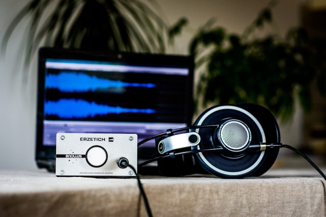 A table holds a silver-metallic headphone amplifier in front of a laptop computer displaying an audio editing software interface with blue audio waveforms on the screen. Large, over-ear headphones with circular ear cups rest near the amplifier, connected by a black cable. In the background, blurred plant foliage adds a natural touch.