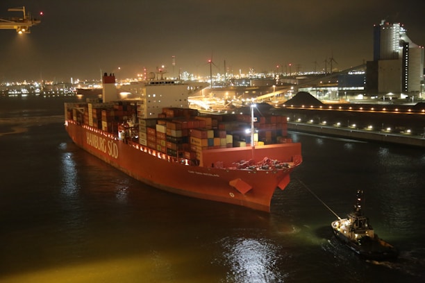 A large container ship with the name Hamburg Süd is being guided by a smaller tugboat in a calm, industrial port at night. The ship is illuminated by lights, and multiple colorful shipping containers are stacked on its deck. In the background, the port is lit with numerous lights under a dark sky, with industrial buildings and cranes visible.