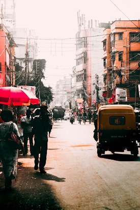 A busy urban street scene with people walking and vehicles such as auto-rickshaws and motorbikes. Street vendors with colorful umbrellas line the sides, and the backdrop consists of multi-storey buildings with various signage. The atmosphere appears bustling and vibrant.