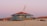A quirky, tilted beach house painted in light blue sits atop sand dunes, with a hammock hanging on its porch. The background features other houses with terracotta roofs and a soft, pinkish-purple sky, suggesting a sunset or sunrise.