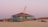 A quirky, tilted beach house painted in light blue sits atop sand dunes, with a hammock hanging on its porch. The background features other houses with terracotta roofs and a soft, pinkish-purple sky, suggesting a sunset or sunrise.
