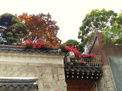 A traditional stone wall with ornate patterns is topped with a line of vibrant red flowers. Behind the wall, trees with lush green and autumnal red leaves add depth and interest to the scene. The wall features a dark tiled roof, and a small lamp is visible near the floral arrangement.