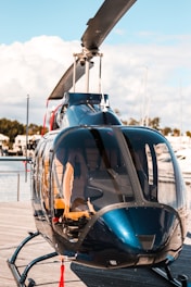 A sleek, glossy helicopter is parked on a wooden platform near a body of water, with calm skies and some clouds in the background. The helicopter's transparent windows reveal its interior seats, and its rotor blades are prominently visible.