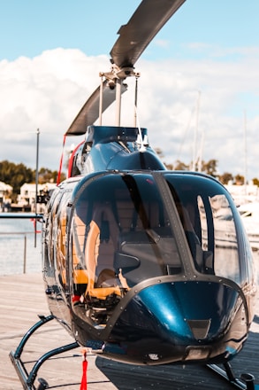 A sleek, glossy helicopter is parked on a wooden platform near a body of water, with calm skies and some clouds in the background. The helicopter's transparent windows reveal its interior seats, and its rotor blades are prominently visible.