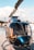 A sleek, glossy helicopter is parked on a wooden platform near a body of water, with calm skies and some clouds in the background. The helicopter's transparent windows reveal its interior seats, and its rotor blades are prominently visible.