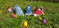 Three colorful sneakers in pastel shades of blue, teal, and red are placed on a grassy field. Surrounding the sneakers are vibrant flowers and Easter eggs in colors such as pink, yellow, and purple, creating a lively and festive scene.