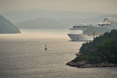 A large cruise ship navigates through a scenic waterway surrounded by lush green hills and rocky shorelines. The sky is overcast, casting a calm and serene atmosphere over the scene. A small lighthouse buoy is visible in the foreground, marking the channel for safe passage.