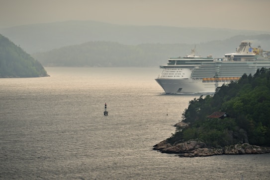 A large cruise ship navigates through a scenic waterway surrounded by lush green hills and rocky shorelines. The sky is overcast, casting a calm and serene atmosphere over the scene. A small lighthouse buoy is visible in the foreground, marking the channel for safe passage.