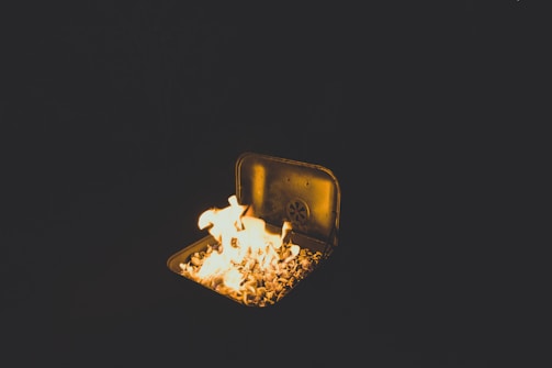 A small fire is burning in a metal container filled with rocks or coals, creating a bright flame against a dark background.