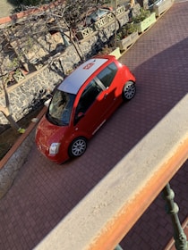 A red compact car parked on a brick driveway with a white roof featuring an orange logo. The surrounding area includes a stone wall and planters with small, leafless trees and some greenery. The driveway is partially shaded by the balcony from which the photo is taken.